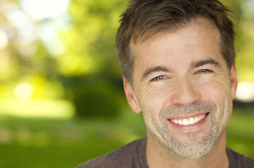 porcelain veneers cosmetic dentistry patient Close-up of a smiling middle-aged man with a bright, confident smile outdoors.