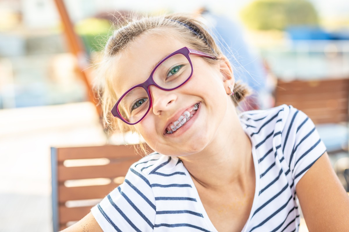 Back to School Braces Colors Smiling girl wearing purple glasses and striped shirt in bright outdoor setting.
