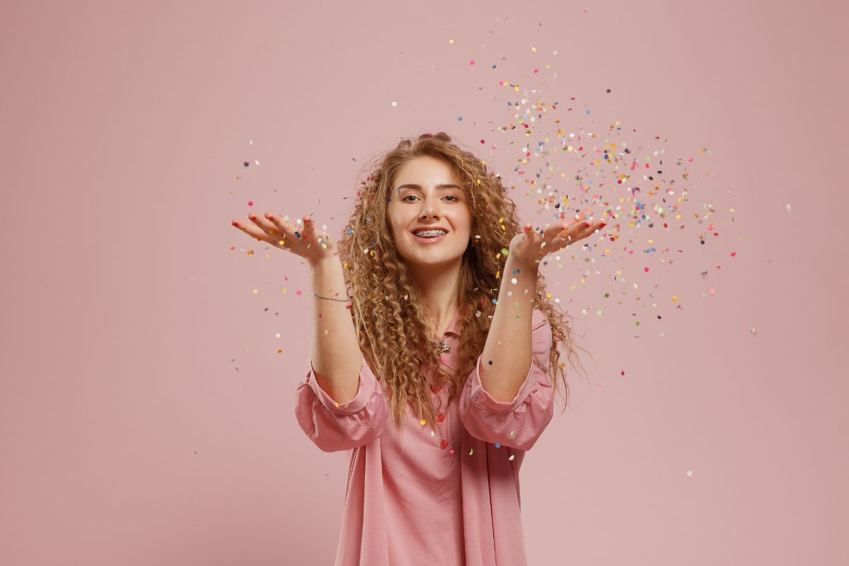 New Braces During the Holidays Happy young woman with curly hair throwing colorful confetti against pink background.