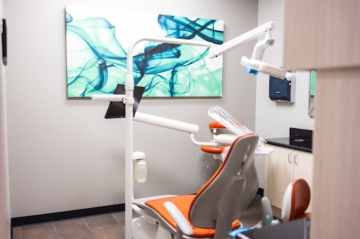 Modern dental operatory with orange and gray patient chair, overhead light, and abstract blue artwork on walls.