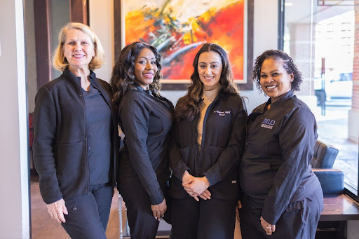Four women in dark professional attire standing together indoors with colorful artwork visible in background.