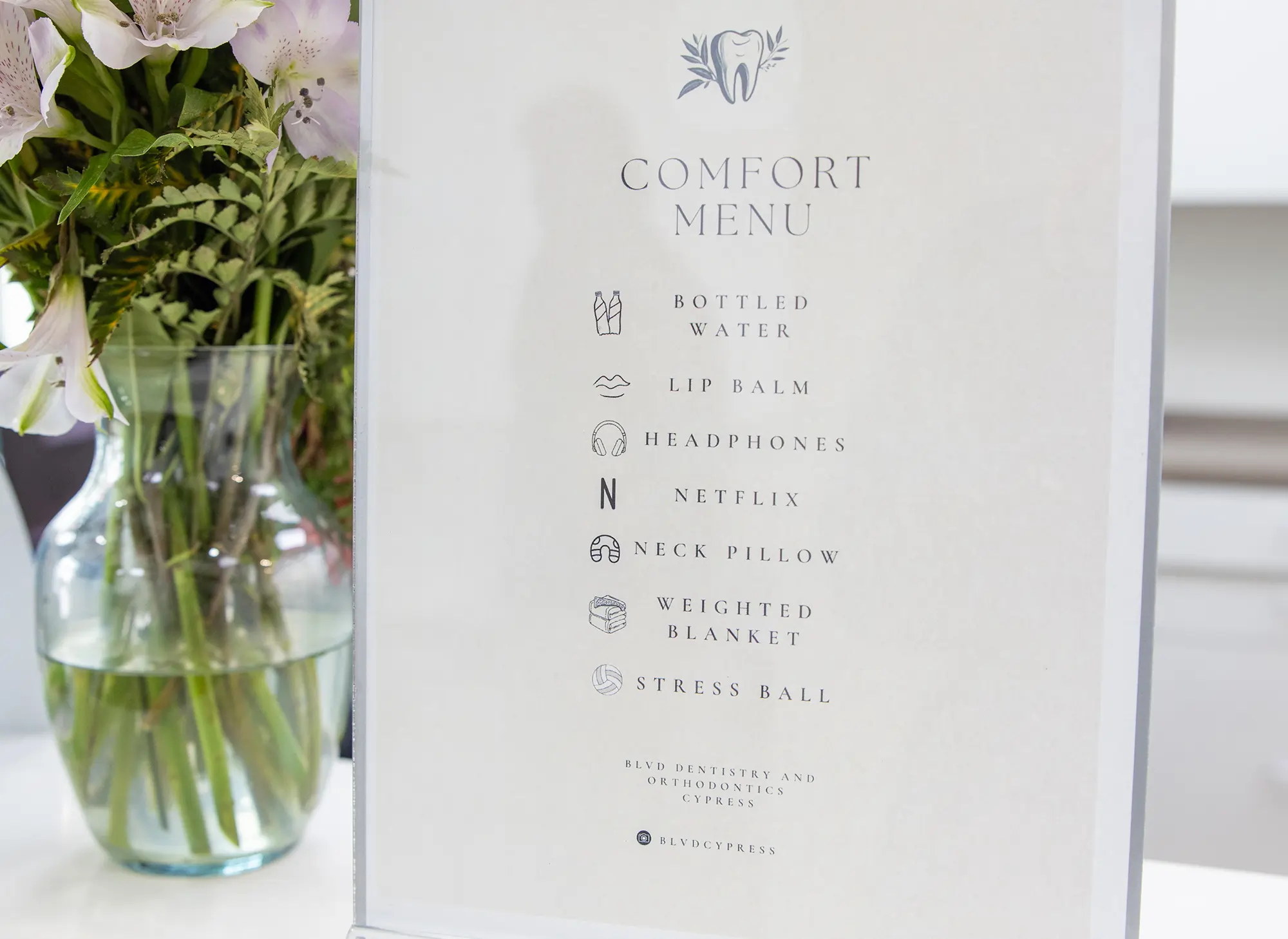 Comfort menu from dental office listing amenities like water, lip balm, headphones, Netflix, neck pillow, weighted blanket, and stress ball next to flowers.