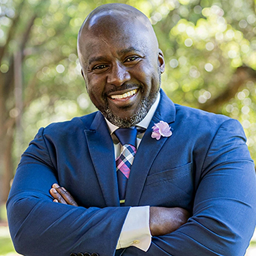 img-dr-Bright Smiling man in blue blazer with arms crossed outdoors among trees.